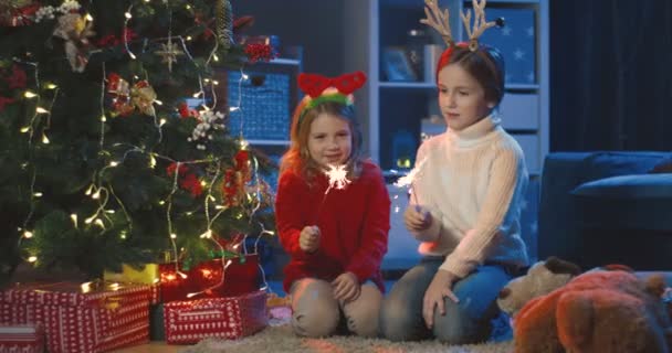 Портретний знімок симпатичних кавказьких маленьких дівчат з кумедними вухами на головах і з блискучими руками сидять на дереві x-mas в прикрашеній кімнаті вночі.. — стокове відео