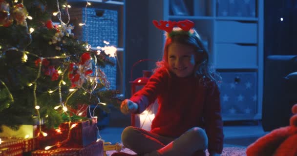 图片说明假日的夜晚，一个可爱的金发白人小女孩坐在圣诞树旁，头上戴着一个闪烁着火花的滑稽耳朵，照片上的她是一个可爱的金发小女孩. — 图库视频影像