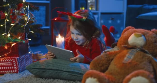 靠近这个快乐的白人小女孩，她躺在沙发上，拿着许多玩具，看着装饰着圣诞夜晚的房间里的平板电脑上有一些有趣的东西. — 图库视频影像