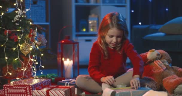 Close up of the joyful small pretty girl unwrapping her x-mas presents while sitting on the Christmas Eve at the tree and smiling. At night. — Stock Video