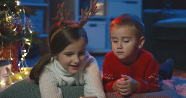 Närbild av de söta små barnen tittar på intressanta och roliga karikatyrer på julafton i det mörka rummet med dekorationer. — Stockvideo