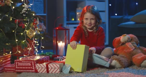Retrato de la linda niña rubia sentada con sus regalos de Navidad y sonriendo a la cámara en el árbol de Navidad en el cuarto oscuro. Cámara alejándose. — Vídeos de Stock