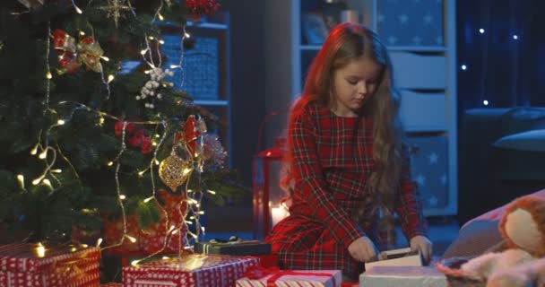 Vit söt liten flicka med långt ljust hår tittar på sina julklappar på den fina vintersemester natt och vara mycket glad. — Stockvideo