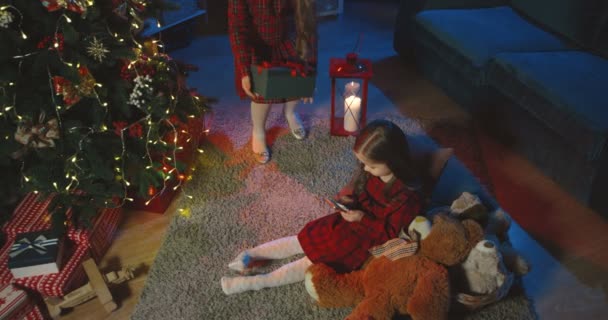 Caucasian cute little girl sitting on the floor with her smartphone in hands in the room on the Chrisrmas Eve, then her friend or sister coming and giving her a present, they hugging. Top view. — Stock Video