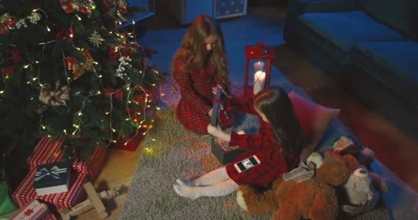 Uitzicht van boven op de twee schattige kleine meisjes zitten op de vloer bij de kerstboom 's nachts en het openen van een doos met cadeautjes - x-mas boom speelgoed. — Stockvideo