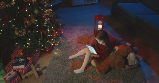Top view on the little cute girl sitting on the floor with tablet computer on the Chrisrmas, then her friend or sister coming and bringing a box with x-mas tree toys. — Stock Video