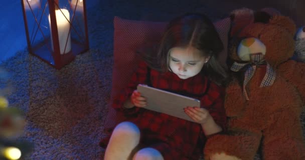 Top tampilan pada gadis kecil yang lucu berbaring di sofa di ruang gelap di pohon x-mas dan menonton sesuatu yang menarik pada perangkat tablet. — Stok Video