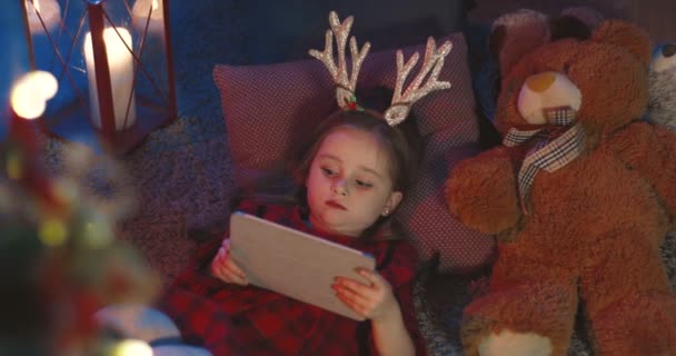 Ovanifrån på den vita lilla söta flicka som ligger på soffan i det mörka rummet på julafton och med hjälp av surfplatta. — Stockvideo