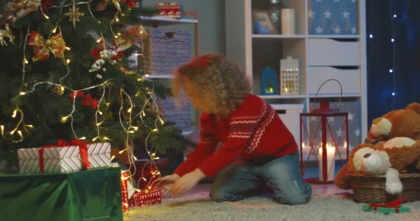 Bouclé petite fille caucasienne assis sous le sapin de Noël dans la chambre joliment décorée et mettre des boîtes avec des cadeaux. — Video