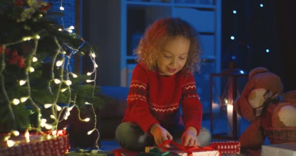 Милая маленькая белая девочка с вьющимися волосами разворачивает рождественскую коробку с подарками на елку ночью. — стоковое видео