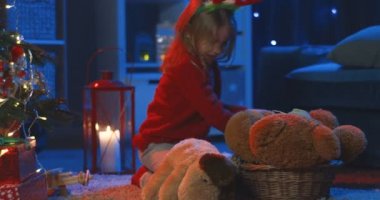 Küçük sevimli beyaz kız, Noel ağacının karanlık odasında otururken oyuncak ayıyla oynuyor..