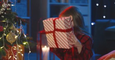 Kafkasyalı küçük şirin kızın portresi büyük Noel hediyesini tutuyor, kutusunu paketliyor ve Noel ağacının karanlık odasında kameraya gülümsüyor. Kapat..