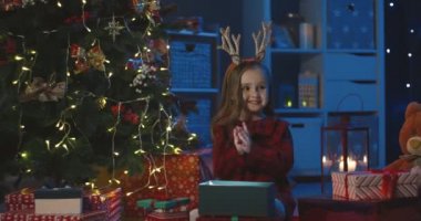 Kafasında komik boynuzları olan sevimli beyaz kız karanlık, rahat bir odada Noel ağacının yanındaki kutuda Noel hediyesine bakıyor..