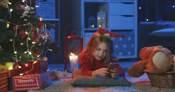 Портрет маленької кавказької дівчини лежить на підлозі з смартфоном і посміхається до камери в темній кімнаті, прикрашеній різдвяним святом.. — стокове відео