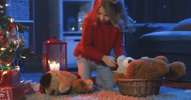 可爱的白种人金发姑娘晚上坐在家里的圣诞树前，把玩具玩具从地板上扔到篮子里. — 图库视频影像