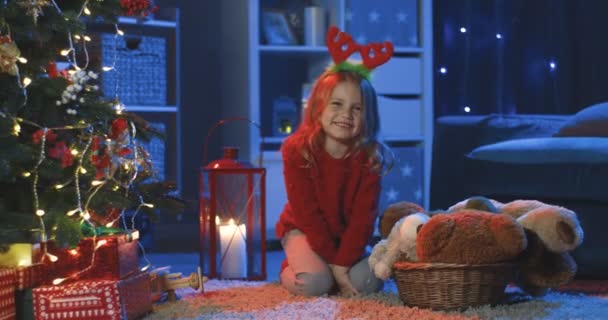 一个可爱的金发白人小女孩坐在圣诞树下的地板上，晚上在黑暗的房间里对着摄像机摆姿势的画像. — 图库视频影像
