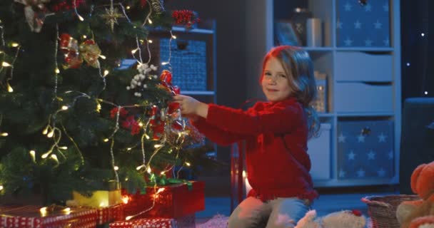 在黑暗的客厅里，一个漂亮可爱的小女孩坐在地板上，装饰着圣诞树，对着摄像机微笑，这是她的画像. — 图库视频影像