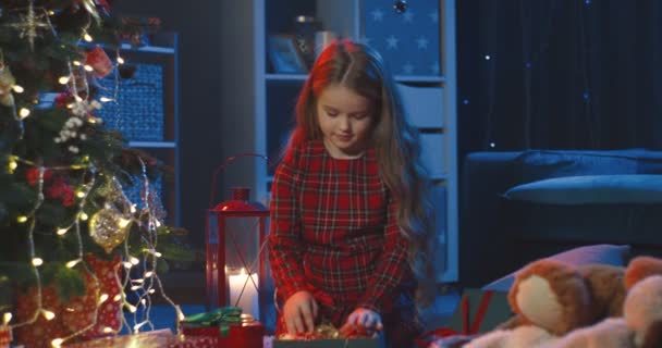 Bella piccola ragazza caucasica con i capelli lunghi seduti nella stanza accogliente buio sul pavimento e decorato albero di Natale. — Video Stock