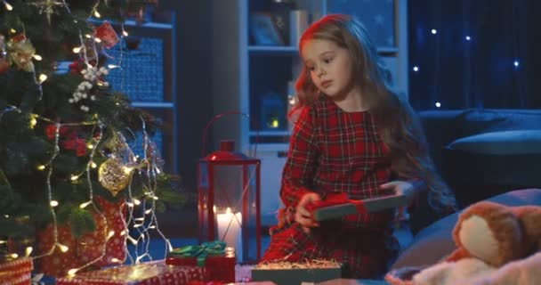 Милая кавказская школьница, сидящая на полу в канун Рождества с рождественскими подарками и кладущая их под елку. — стоковое видео