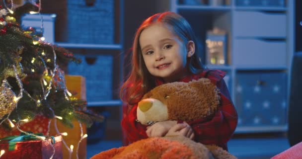 Nahaufnahme des hübschen kleinen Mädchens, das auf dem Boden sitzt, Teddybär umarmt und im dunklen Wohnzimmer am Weihnachtsbaum in die Kamera lächelt. Porträt. — Stockvideo