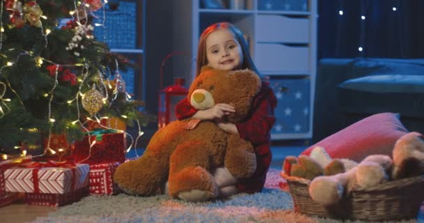 Porträtt av den söta och lilla flickan som sitter på golvet, kramar nalle och ler mot kameran i det mörka rummet vid julgranen på julafton. — Stockvideo