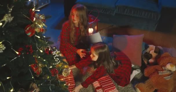 View from above on the two cute Caucasian small girls sitting at the x-mas tree with the presents in the living room in the evening. — Stock Video