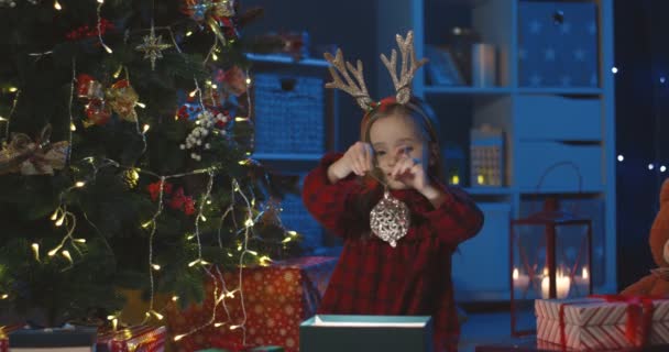 Маленька кавказька дівчинка з кумедними рогами на голові бере іграшковий м'яч з коробки і прикрашає дерево Х-мас.. — стокове відео