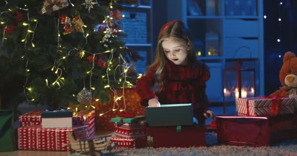 Hübsches kaukasisches kleines Mädchen holt ein Spielzeughirsch aus der Schachtel und schmückt den Weihnachtsbaum damit abends in der gemütlichen dunklen Stube. — Stockvideo