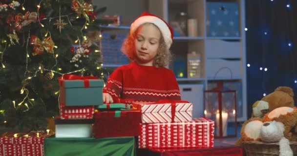 Портрет милого белого маленького ребенка в шляпе Санта-Клауса, улыбающегося со всеми ее рождественскими подарками на елку. — стоковое видео