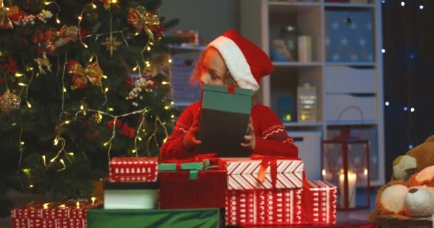 Belle petite fille caucasienne dans le chapeau du Père Noël prenant boîte cadeau x-mas, étreignant et écoutant tout en prenant à son oreille. À la maison avec un sapin de Noël. — Video