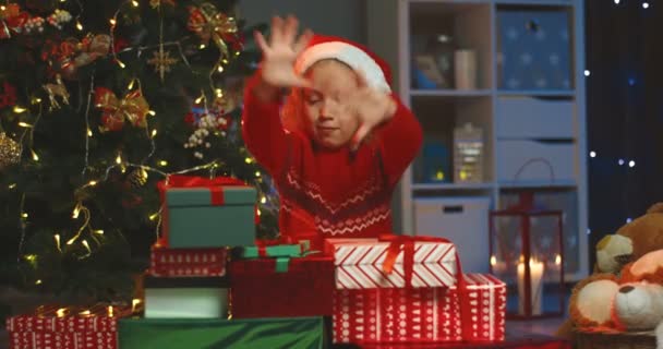 Портрет веселого симпатичного белого малыша в шляпе Санта Клауса, улыбающегося в камеру, обнимающего все рождественские подарки на рождественской елке дома. — стоковое видео