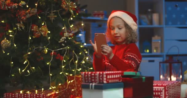 Zdjęcie portretowe uroczego białego dziecka w kapeluszu Świętego Mikołaja stukającego i oglądającego coś na smartfonie z zaskoczoną twarzą na drzewie X-mas. — Wideo stockowe