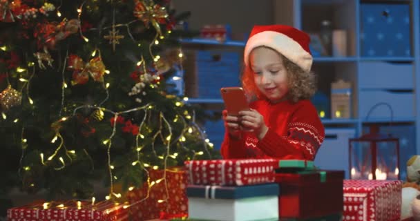 Belle petite fille caucasienne dans le chapeau du Père Noël jouer à un jeu sur le smartphone joyeusement. À la maison avec un sapin de Noël. — Video