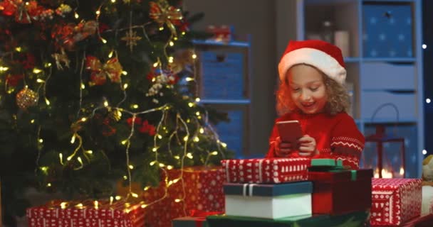 Porträt des fröhlichen hübschen kaukasischen Kindes im Hut des Weihnachtsmannes, das sein Smartphone benutzt und zu Hause am Weihnachtsbaum ein Spiel darauf spielt. — Stockvideo