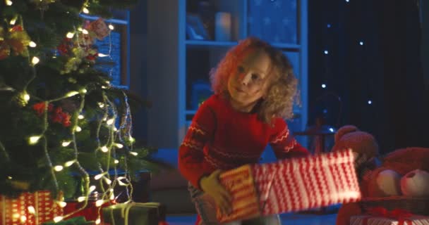 Portrait de la mignonne fille bouclée ouvrant une grande boîte avec cadeau de Noël au sapin de Noël la nuit. — Video