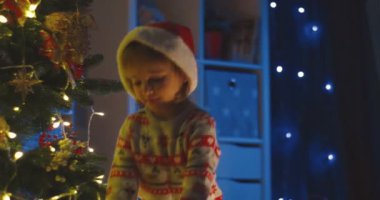 Beyaz çocuk Noel Baba şapkası takıyor ve Noel ağacında Noel hediyesi veriyor..