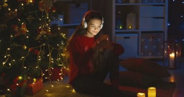 Kafkasyalı genç güzel kız kulaklıklı müzik dinliyor ve yılbaşı ağacının karanlık odasındaki akıllı telefona dokunuyor..