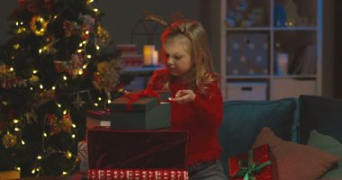 Kafasında komik boynuzları olan güzel beyaz bir kız Noel ağacının altında hediyelere bakıyor ve içeriden aydınlatan bir kutu açıyor..