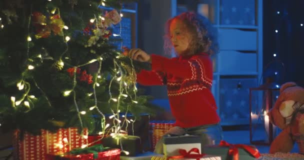 Mignon caucasien petit bouclé enfant décoration arbre de Noël avec jouets ballons dans la chambre noire dans la salle du soir. — Video