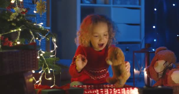Härlig vit lockig liten unge öppna en låda med sin julklapp från Santa och ta ut leksaker på julgran på natten. — Stockvideo