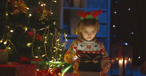 Portrait de la petite caucasienne mignonne avec une corne de cerf sur la tête ouvrant une boîte avec cadeau de Noël lors de la nuit de Noël magique. — Video