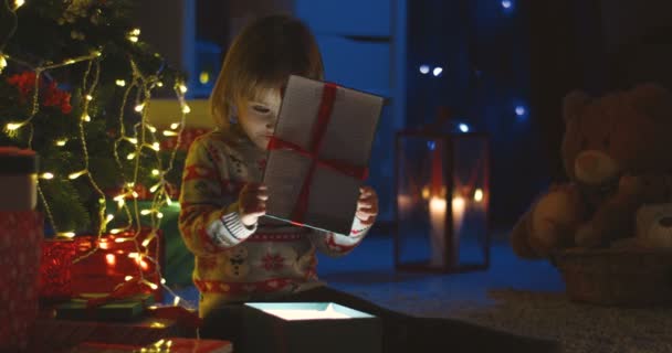 Ritratto della graziosa piccola ragazza caucasica che apre una scatola regalo x-mas e guarda dentro mentre brilla la notte di Natale. — Video Stock