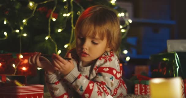 Mignon petit garçon caucasien couché au sapin de Noël parmi les cadeaux de Noël avec un crayon à la main dans la soirée. — Video