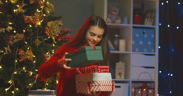 Retrato de la muchacha bonita adolescente caucásica sonriendo a la cámara y sosteniendo muchas cajas con regalos en la acogedora habitación en el árbol de x-mas. — Vídeo de stock