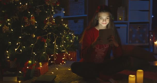 Retrato de la hermosa adolescente caucásica usando un dispositivo de tableta mientras ve algo y golpea en el árbol de Navidad por la noche. — Vídeo de stock