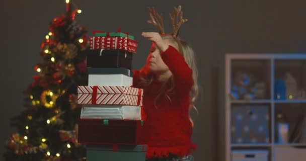Pretty small Caucasian cute girl in the funny horns on her head taking her x-mas present from the top and running away at the Christmas tree. — Stock Video