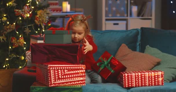 Blank klein schattig tiener meisje dragen grappig hert hoorns op zoek naar kerstcadeaus terwijl zitten op de bank in de woonkamer bij de x-mas boom. — Stockvideo