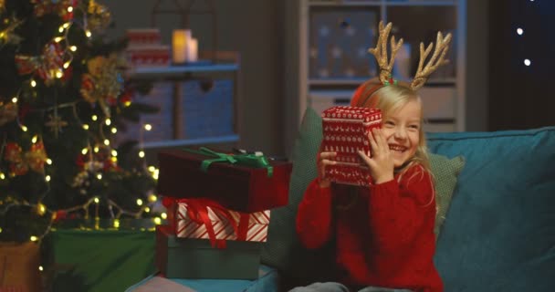 Portrait de la jolie fille blanche blonde avec de drôles de cornes de cerf sur la tête assise sur le canapé à l'arbre de Noël et heureuse avec son cadeau du Père Noël. — Video