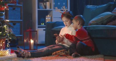 Beyaz tenli genç kız akıllı telefonda oynuyor ve küçük kardeşi de Noel ağacında yerde otururken onu izliyor..