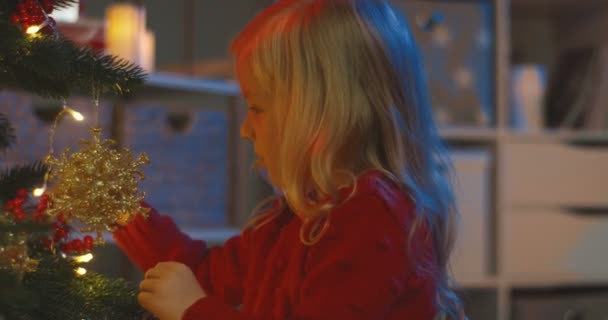 Close up of the cute blonde Caucasian little girl playing with decorating toys from the Christmas tree in the dark room. — Stock Video